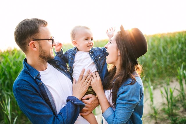 Young hipster father in glasses, mother holding cute baby in summer cornfield